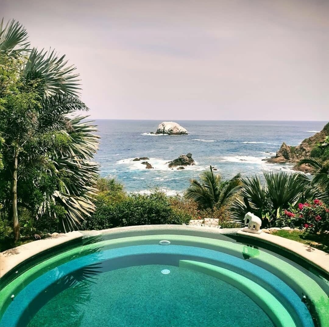 An inviting circular pool with turquoise water is surrounded by lush tropical plants. The ocean view features a rocky outcrop in the distance, with waves gently crashing on the shore, creating a serene coastal atmosphere.