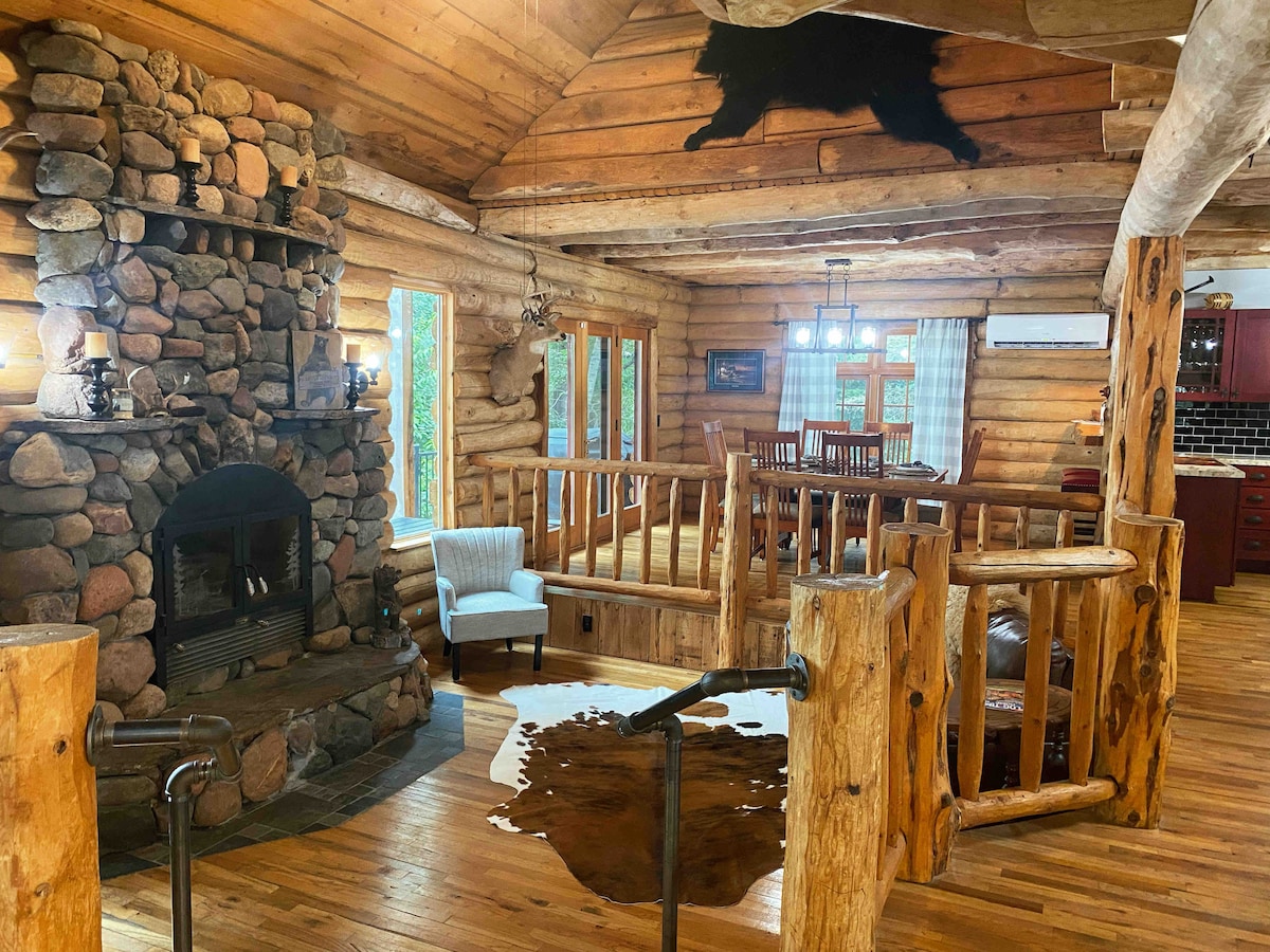 The main living area features a large stone wood-burning fireplace as a focal point, surrounded by rustic wooden beams and log walls. A cozy seating area is arranged beside the fireplace, and natural light enters through multiple large windows, enhancing the inviting environment.