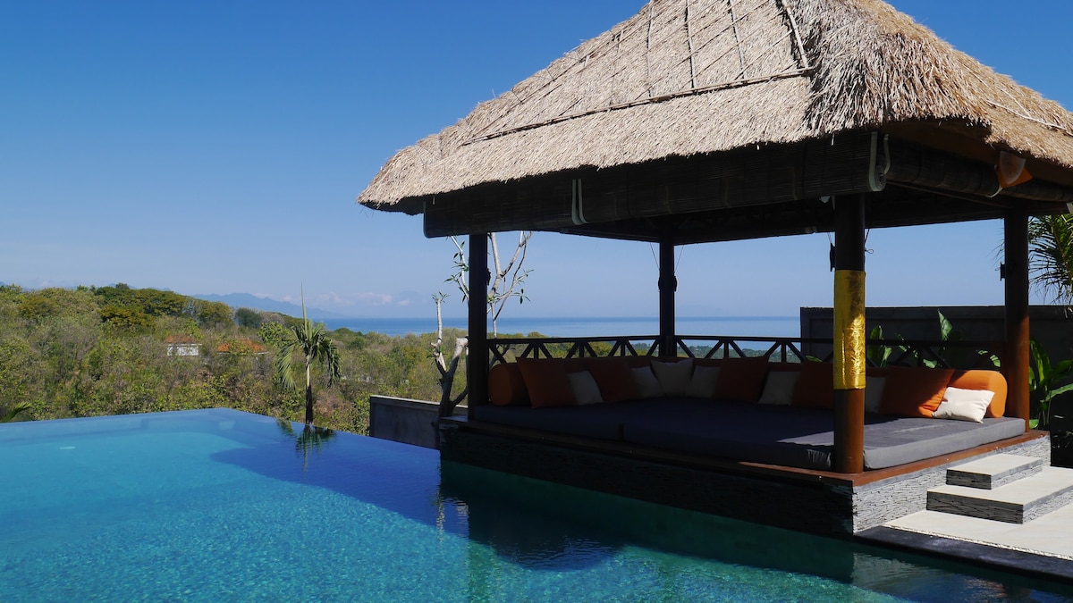 A traditional gazebo with a thatched roof overlooks a tranquil infinity pool, blending seamlessly with the clear blue sky. Cozy cushions line the seating area, inviting relaxation while providing an unobstructed view of the lush surroundings and the ocean beyond.