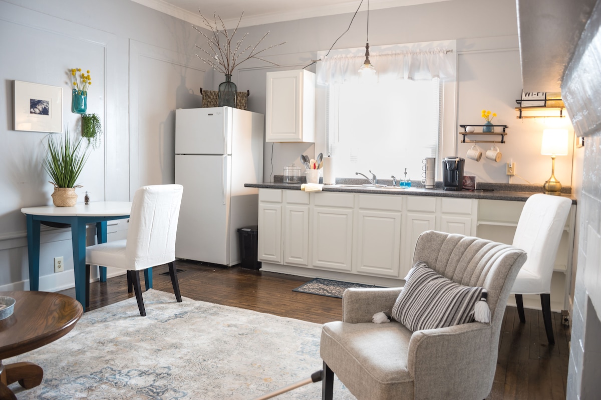 A well-lit kitchen and living area features white cabinetry and a full-sized refrigerator. A comfortable seating area includes a gray armchair and a round dining table with two chairs. Natural light pours in through a large window, enhancing the cozy atmosphere.