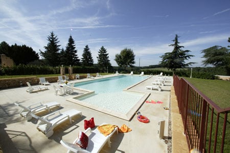 A spacious outdoor pool area features a large swimming pool surrounded by sun loungers. Lush green trees provide shade, while a well-maintained lawn borders the space. Colorful pool accessories are scattered across the deck, enhancing the inviting atmosphere of this tranquil setting.