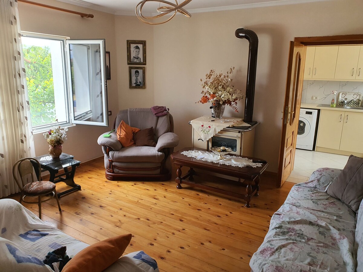 A cozy living room features a light wooden floor and natural light streaming through a large window. Seating options include a plush armchair and a floral-patterned sofa. A wooden coffee table occupies the center, and a small decorative table holds a flower arrangement.