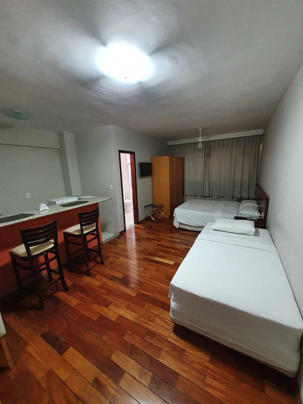 A well-lit room features wooden flooring and a mix of furnishings. A spacious bed is positioned beside a single bed. Two barstools face a kitchen counter, and a wardrobe offers additional storage. Natural light enters through the sheer curtains of a nearby window.