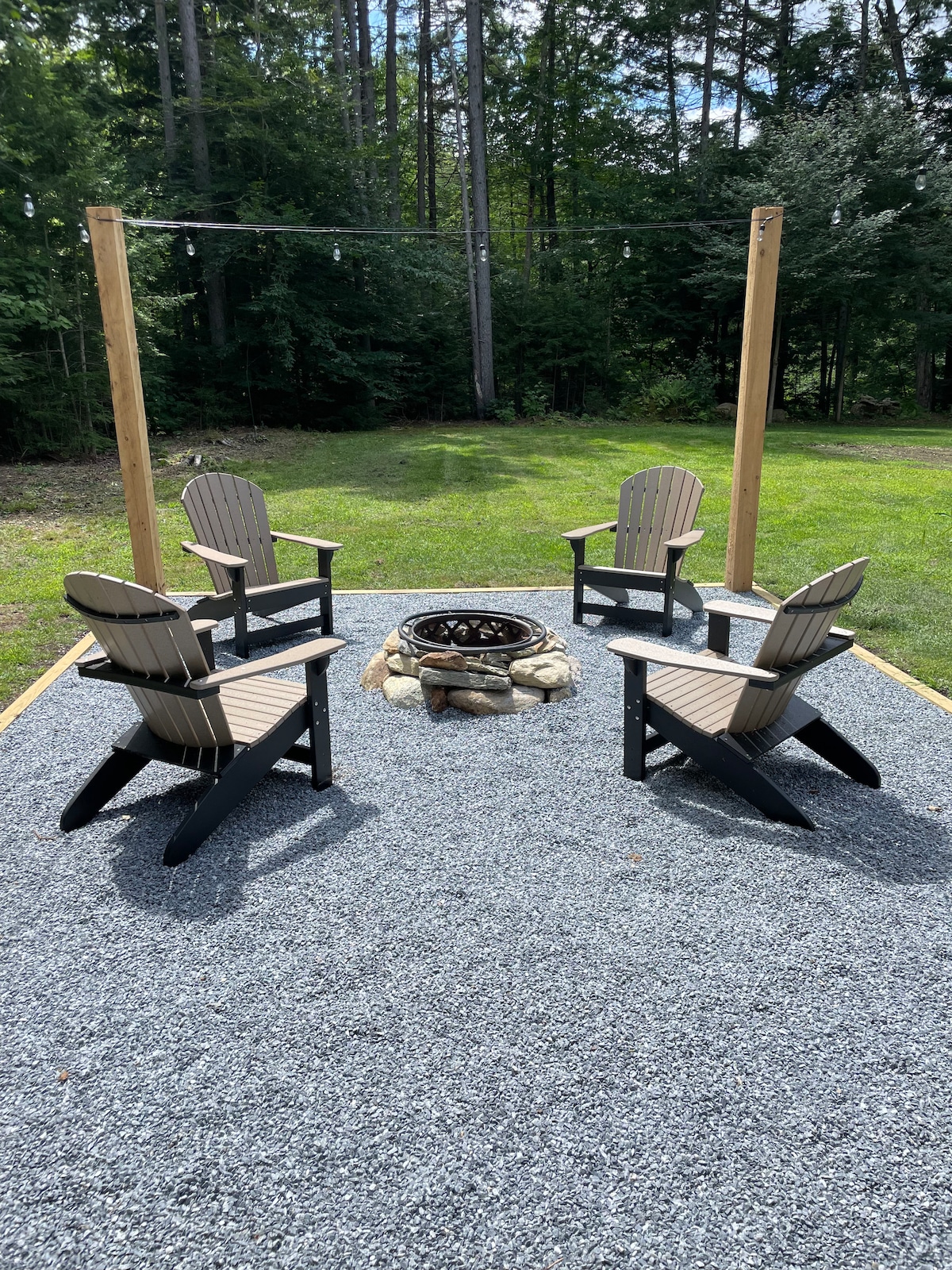 An outdoor fire pit area is surrounded by four Adirondack chairs, positioned on a gravel surface. The setup is bordered by trees, creating a serene environment. Soft lighting can be seen hanging overhead, enhancing the setting for gatherings around the fire.