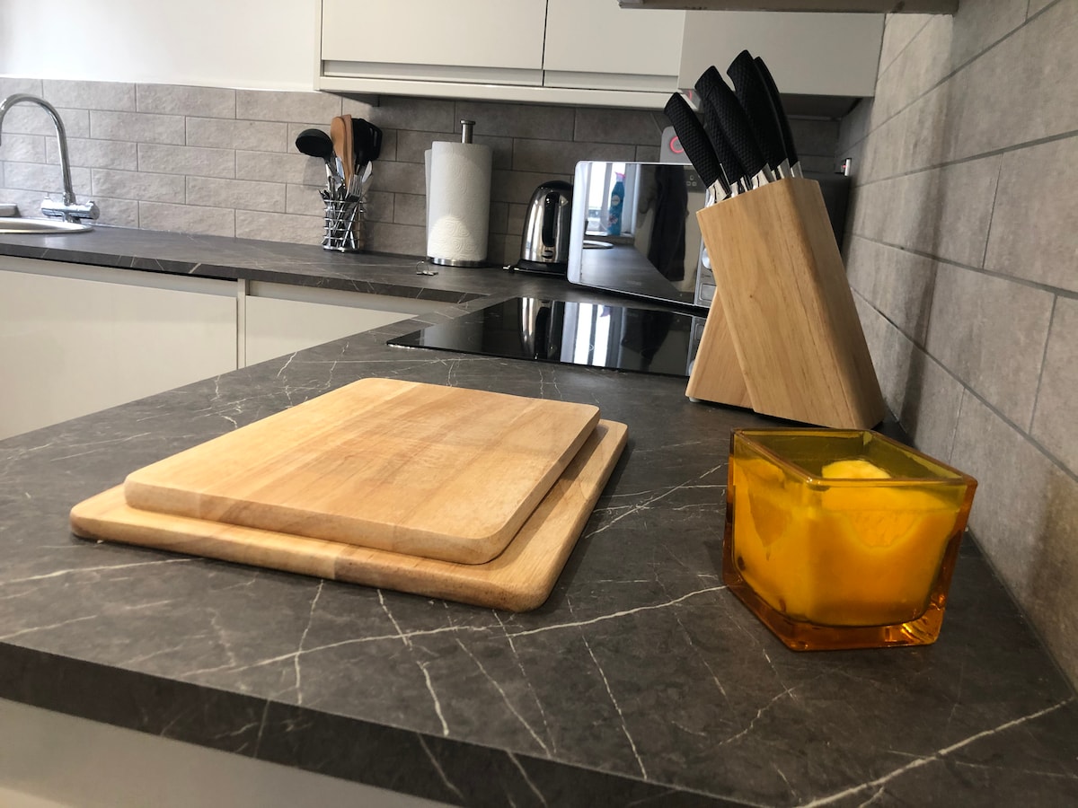 The modern kitchen countertop features a wooden cutting board and an amber-colored container. A set of knives is neatly arranged in a wooden holder, alongside a kettle and a microwave, all placed against a stylish grey tiled backsplash.