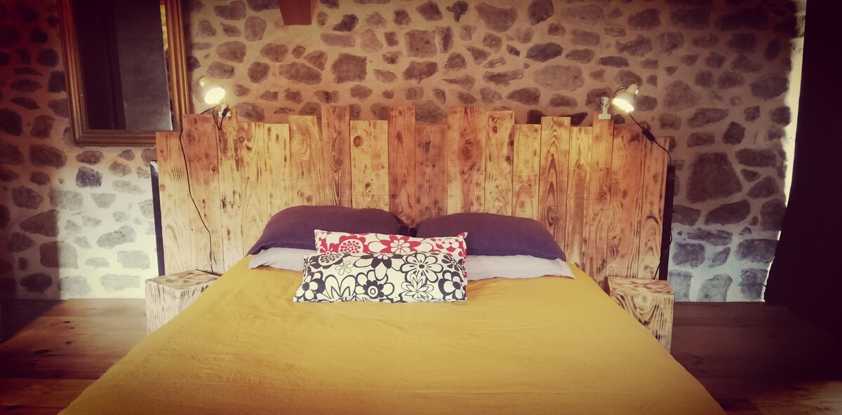 A spacious bed is framed by a rustic wooden headboard made from reclaimed wood. Two bedside lamps provide soft lighting, and decorative pillows add a touch of color. The stone wall enhances the natural feel of the room.