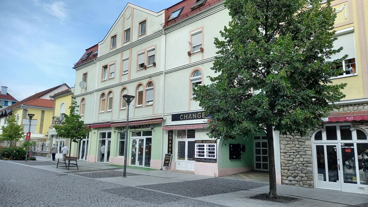 A street view showcases a charming building with a pastel-colored facade, featuring multiple windows and awnings. Beneath a clear blue sky, a tree provides shade in front of the structure, while pedestrian footpaths invite leisurely strolls.
