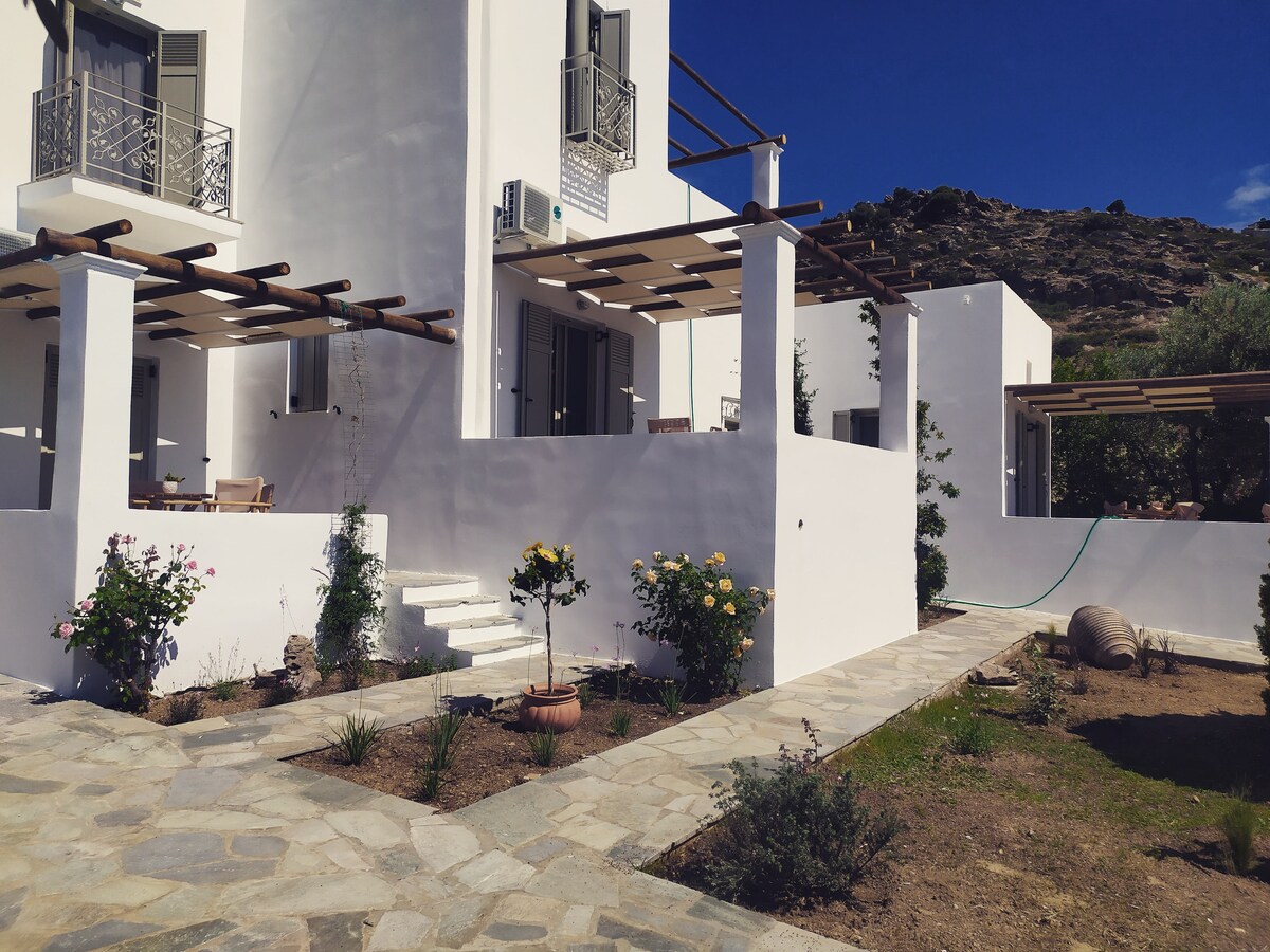 A bright, white building is positioned under a clear blue sky, featuring a covered porch with wooden beams. A stone pathway leads through a landscaped area with plants and flowers, showcasing the inviting outdoor space.