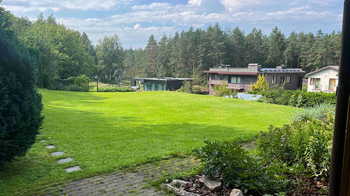A spacious green lawn extends towards the edge of a serene forest, featuring well-maintained landscaping. Various trees and shrubs are present, with a clear view of neighboring homes in the background. The sky appears bright and partly cloudy.