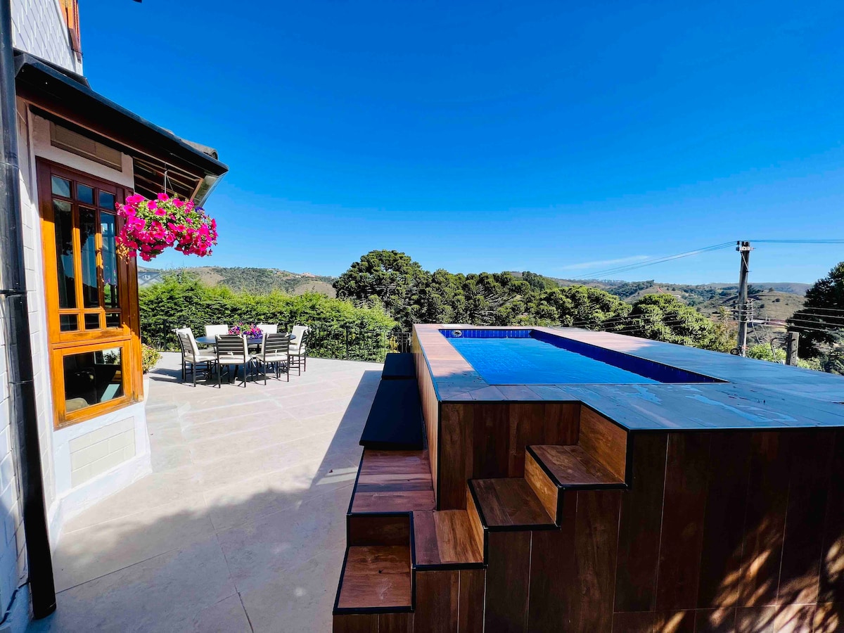 A modern swimming pool is featured on an outdoor patio, surrounded by natural views. Steps lead into the pool area, which is bordered by wooden decking. A stylish table with chairs is positioned nearby, ideal for dining outdoors. Bright flowers add a touch of color.