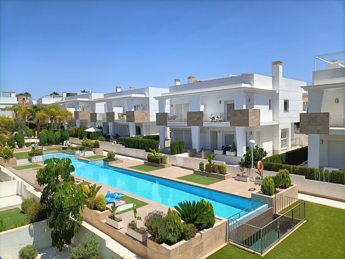 A modern villa complex is shown with two stories and a large communal pool in the center. Lush greenery and neatly manicured lawns surround the pool area, providing a serene outdoor environment. Ample space for relaxation and leisure activities is available.
