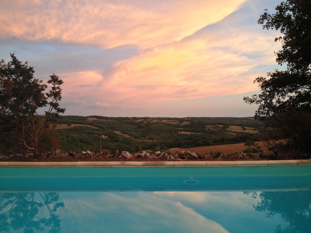 A serene pool reflects the colors of a sunset, surrounded by lush greenery and distant rolling hills. The sky displays soft hues of pink and purple, creating a tranquil backdrop for relaxation.
