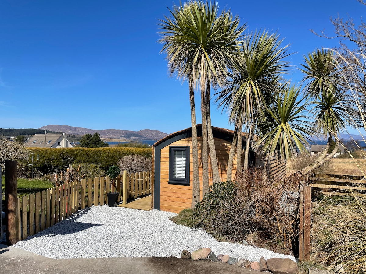 A wooden pod is surrounded by lush palm trees and gravel pathways. The clear blue sky highlights the structure's natural setting, with distant hills visible beyond the greenery. The entrance is framed by a wooden fence, providing a welcoming approach.