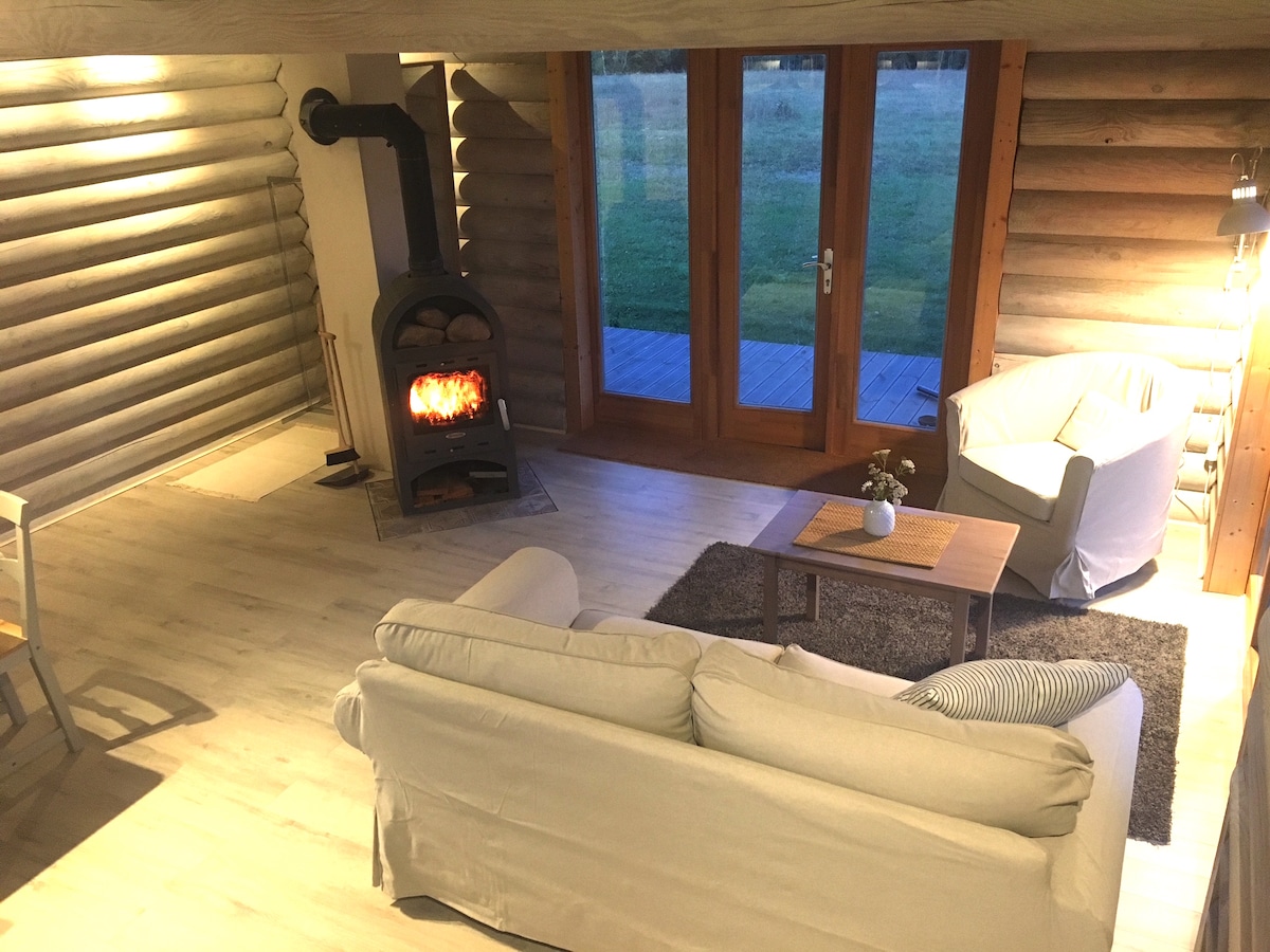 A cozy interior showcases a comfortable sofa and an armchair arranged around a wooden coffee table. A modern wood stove adds warmth to the space, which features large glass doors that provide a view of the surrounding nature.