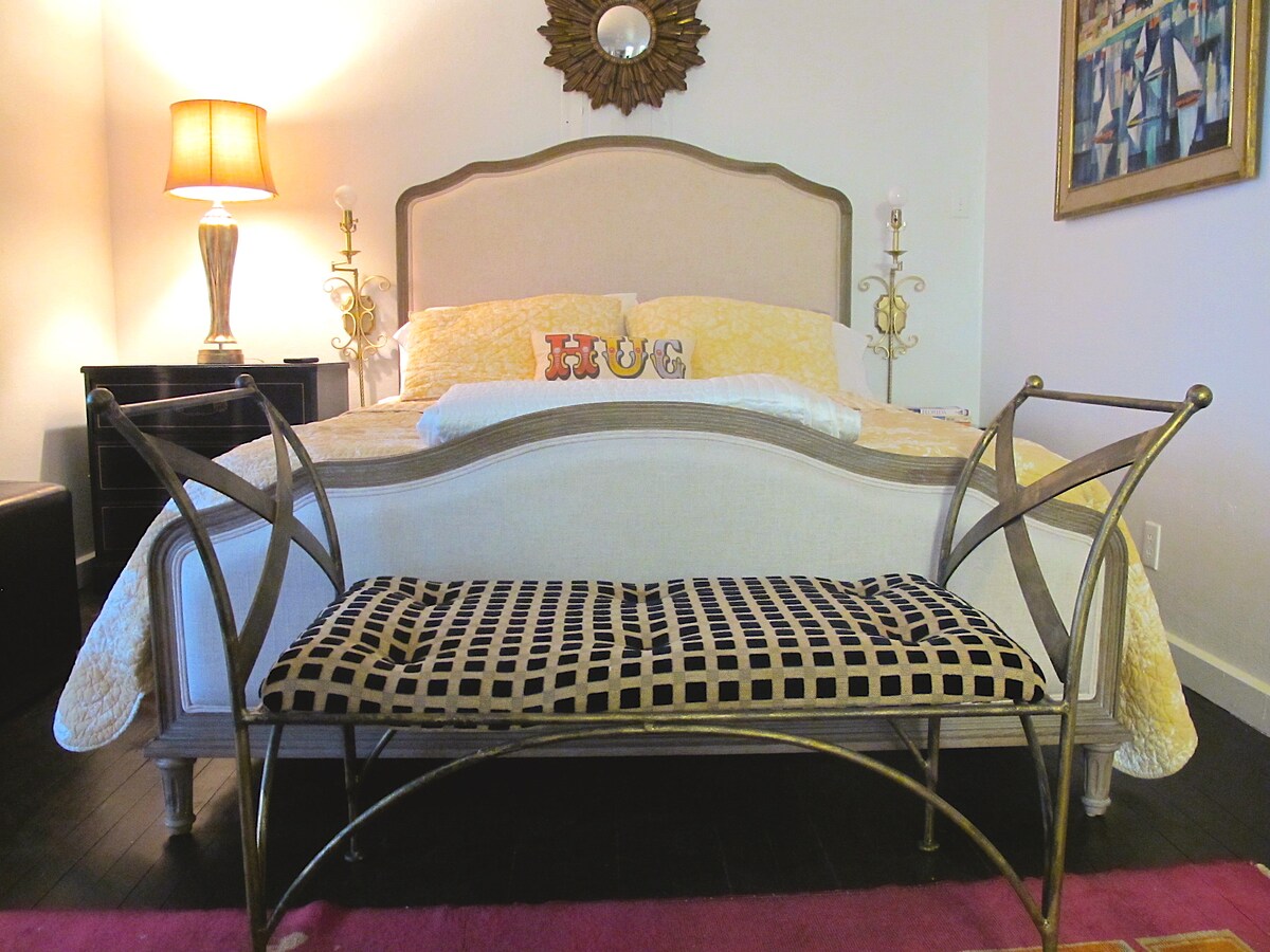A neatly arranged sleeping area features a queen-sized adjustable bed dressed in a yellow quilt. A decorative bench with a grid pattern stands at the foot of the bed, and warm light emanates from a lamp beside the bed. A mirror and framed artwork add visual interest to the space.