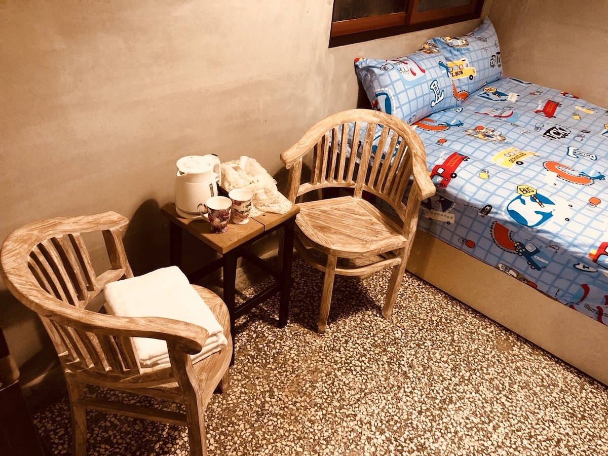 The room features a cozy sleeping area with a bed adorned in a colorful car-themed blanket. Two wooden chairs are positioned beside a small table, which holds a kettle and cups, creating a welcoming space for relaxation.