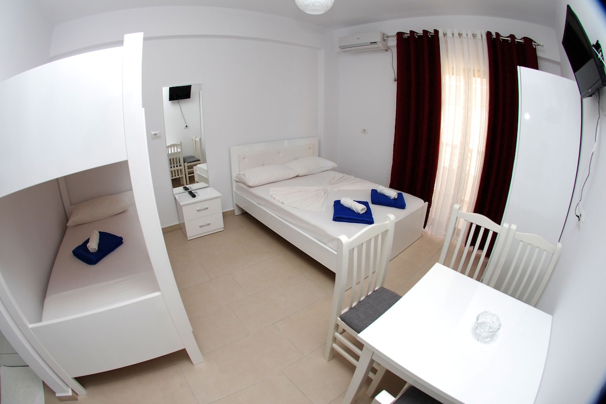 A bright sleeping area features a spacious bed and a bunk bed, all in a modern white design. A small dining table with two chairs is positioned nearby, and a large window with sheer curtains allows natural light to illuminate the room. A TV is mounted on the wall.