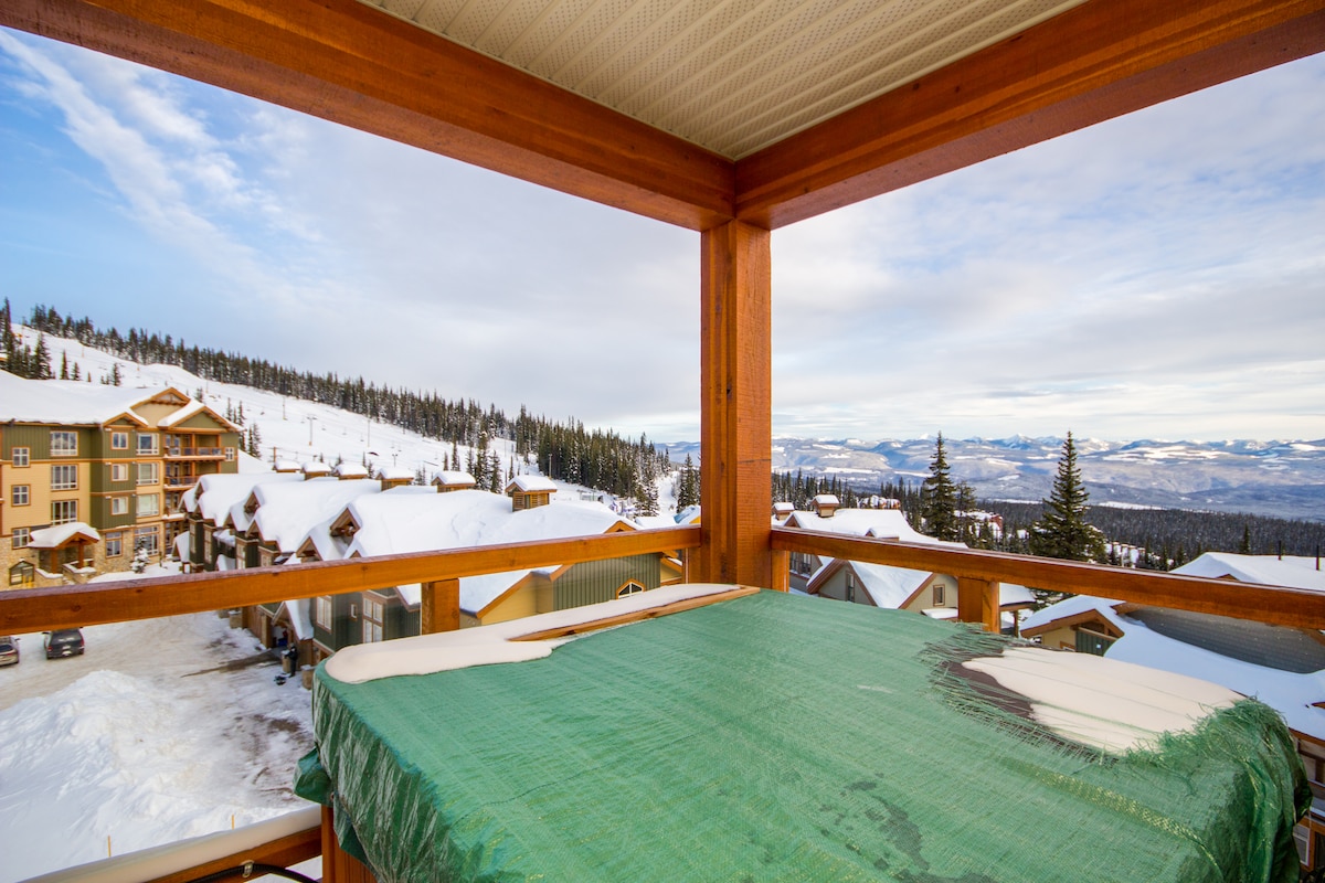 A private hot tub is situated on a wooden deck, overlooking a winter landscape filled with snow-covered buildings and trees. The horizon reveals distant mountains under a partly cloudy sky, providing a scenic backdrop for outdoor relaxation.