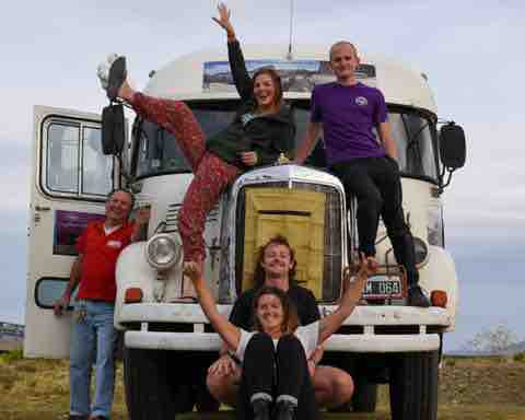 A group of four individuals poses playfully in front of a vintage motorhome. The vehicle features a distinct front grille and large headlights. The backdrop consists of an open landscape under a cloudy sky, contributing to a sense of adventure.