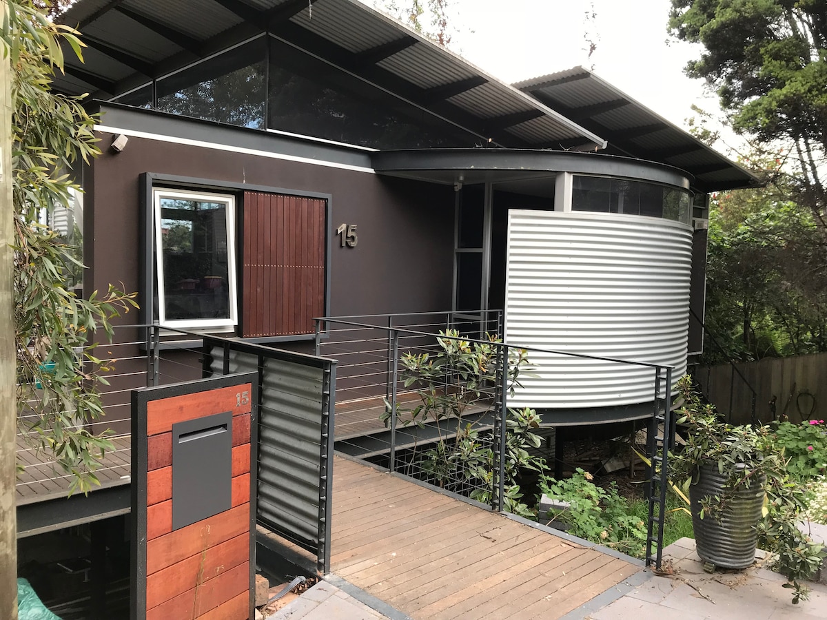 The modern apartment features a unique architectural design with a curved exterior and large windows. A wooden deck leads to the entrance, surrounded by greenery, enhancing the connection with nature. The mailbox is visible near the entrance, indicating the apartment's private access.