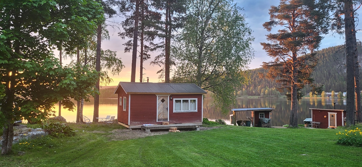 The small red cabin is set amidst tall trees, surrounded by lush green grass, with a peaceful river reflected in the background. A simple porch leads to the cabin door, while additional service buildings are visible nearby.