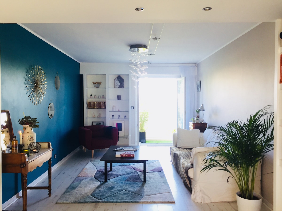 A spacious living room is presented, featuring a combination of blue and neutral walls. A round coffee table sits on a patterned rug, with a comfortable sofa and red armchair nearby. Natural light floods the area, enhancing the greenery from a potted plant.