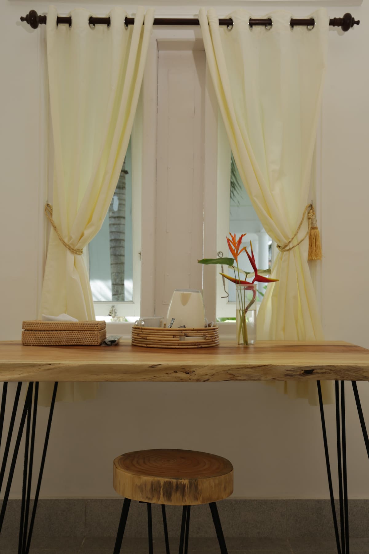 A wooden table with slender black legs is positioned against a window framed by light yellow curtains. A small stool complements the table. A fresh flower arrangement sits on the table alongside a woven basket and tea-making facilities, providing a serene workspace.