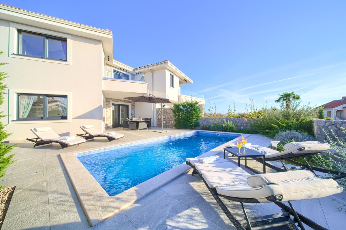 A private swimming pool is surrounded by lounge chairs and tables, emphasizing a relaxing outdoor space. Lush greenery borders the area, and a portion of the house is visible in the background, providing an inviting setting for leisure and enjoyment.