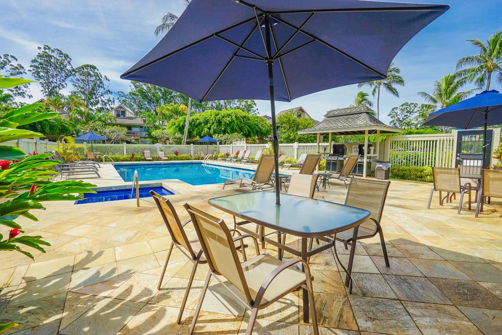 A spacious pool area is shown, featuring a clear blue pool surrounded by lounge chairs and tables with umbrellas. Lush greenery and palm trees add to the tropical ambiance, while shaded seating offers a comfortable spot for relaxation.