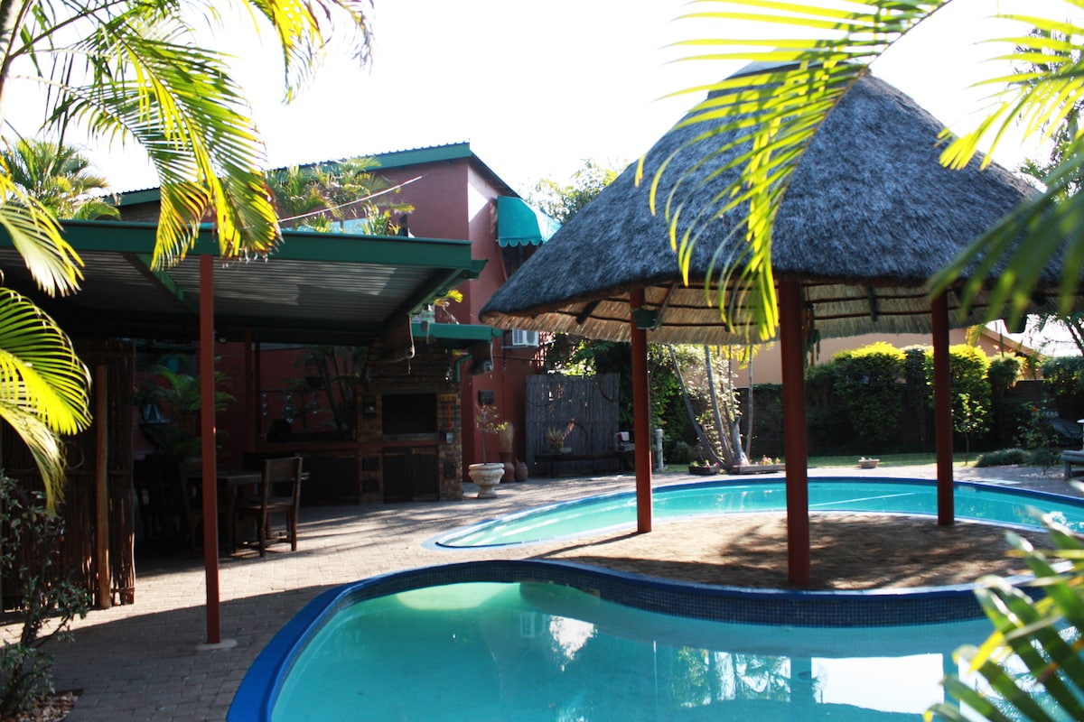 A tranquil swimming pool is nestled beside a thatched-roof gazebo. Surrounding palm trees create a lush atmosphere, while an outdoor dining area and a built-in braai are visible under the covered patio in the background.