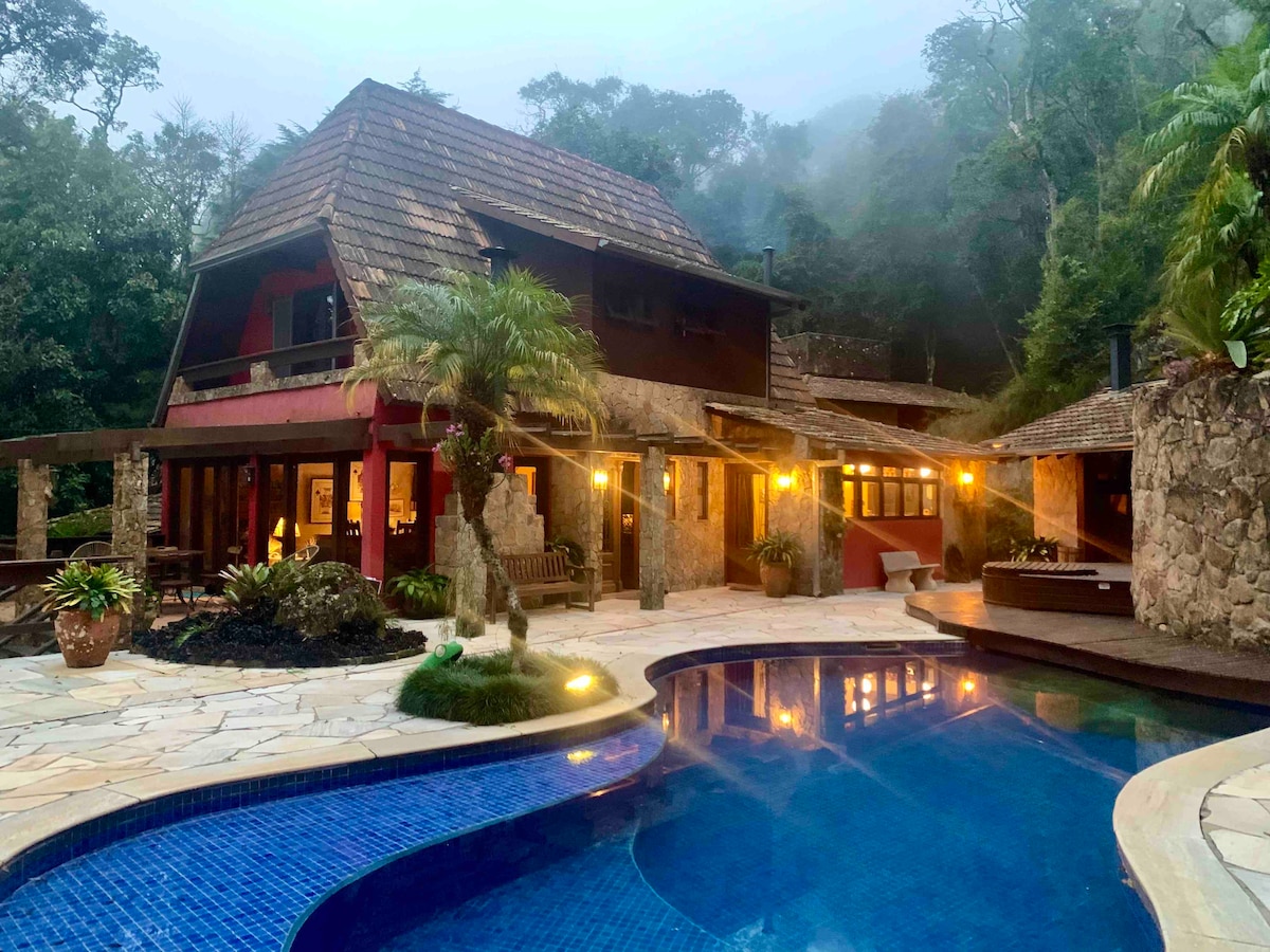A charming mountain house is set among lush greenery, featuring a stone facade and a rustic roof. A pool with blue tiles reflects the surrounding landscape. Warm lights illuminate the exterior, creating a welcoming atmosphere in the early evening.