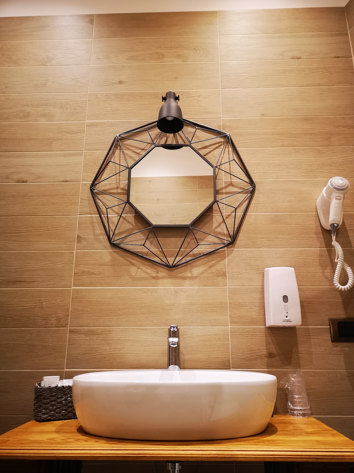 The bathroom features a modern basin with a sleek, rounded design situated on a wooden countertop. A geometric mirror is mounted above the sink, reflecting the contemporary decor. The wall is adorned with wooden panels, and a stylish wall sconce provides illumination.