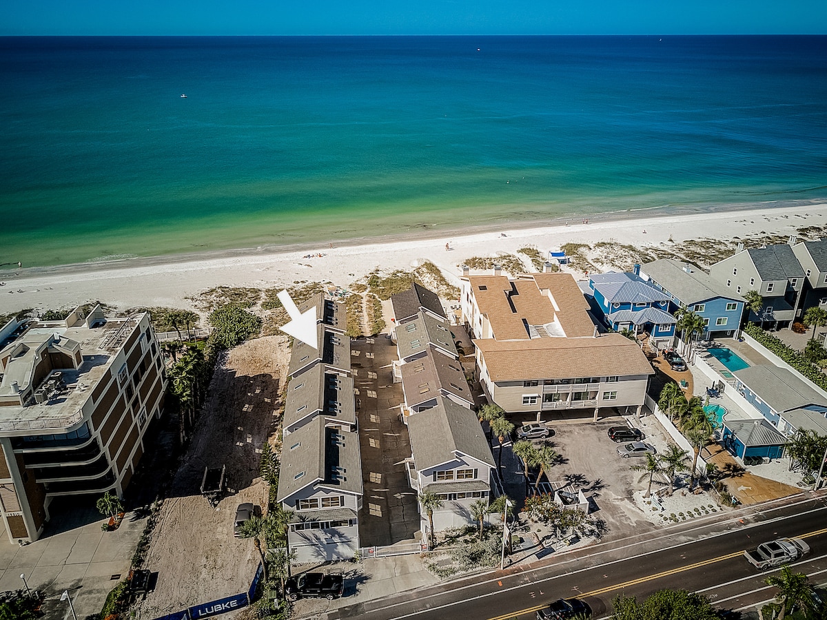 An aerial view captures a row of beach homes adjacent to a private beach. The sandy shore and gentle waves create a serene backdrop. The clear blue water contrasts with the white sand, highlighting the tranquility of the coastal setting.