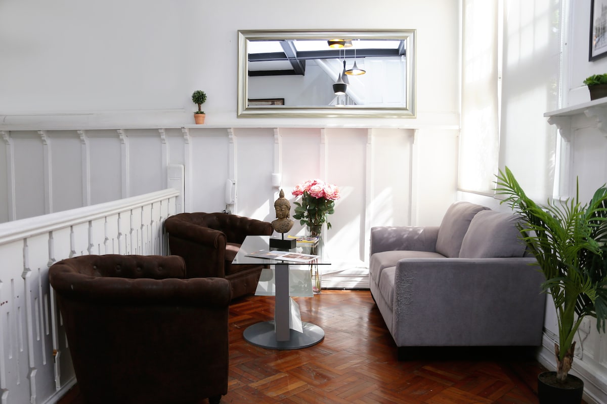A cozy seating area features two comfortable armchairs and a modern sofa arranged around a small glass table. Natural light enters through large windows, illuminating plants and a decorative vase with flowers. A large mirror enhances the sense of space.