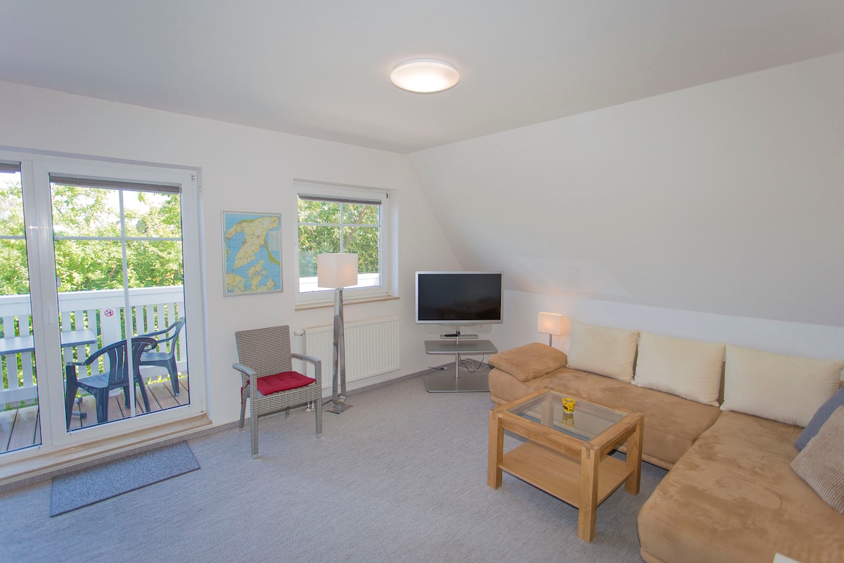 The living area features a corner couch and a wooden coffee table. A television is positioned near a floor lamp, providing soft lighting. Two windows allow natural light to fill the space, leading to a balcony with outdoor seating.