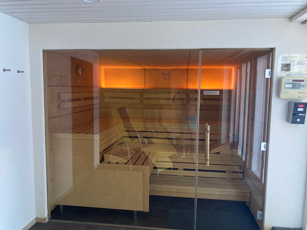 A modern sauna is featured, enclosed by glass walls that allow for visibility. Wooden benches line the interior, with a subtle ambient light creating a calming atmosphere. A clock is mounted on the wall, providing convenience for guests. A door leads to the entrance.