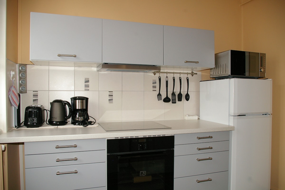 A functional kitchen area is presented, featuring modern cabinetry in light grey. Appliances include a refrigerator, a microwave, a coffee maker, and a kettle. A stovetop is installed on the counter, accompanied by hanging utensils. Ample storage space is offered within the cabinets.
