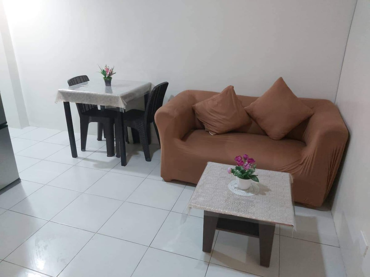 A simple living area features a brown sofa with two decorative cushions, paired with a dining table set for four. A small table holds a flower arrangement, while light-colored tiles create a bright and open atmosphere.