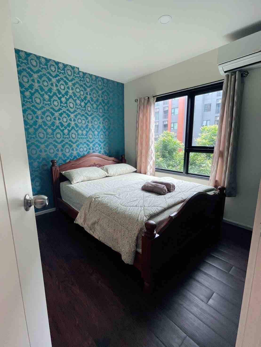 A cozy bedroom features a wooden bed with a light-toned quilt, positioned near a large window dressed with sheer curtains. The vibrant blue patterned wall adds visual interest, while natural light filters into the space, creating a welcoming environment.