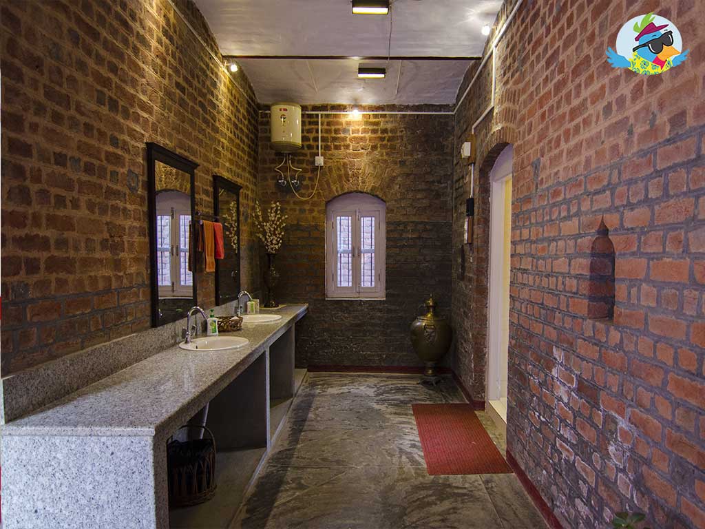 A spacious bathroom features exposed brick walls that provide a rustic touch. Two large sinks are placed on a long granite countertop, complemented by two mirrors. Natural light filters in from a small window, with a traditional clay pot positioned against the wall.