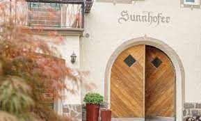 An entrance is marked by an arched wooden door with diamond-shaped panels. The building features a warm, textured facade, complemented by decorative signage reading 'Sunnhofer.' Potted greenery is positioned on either side, adding a touch of nature to the entryway.
