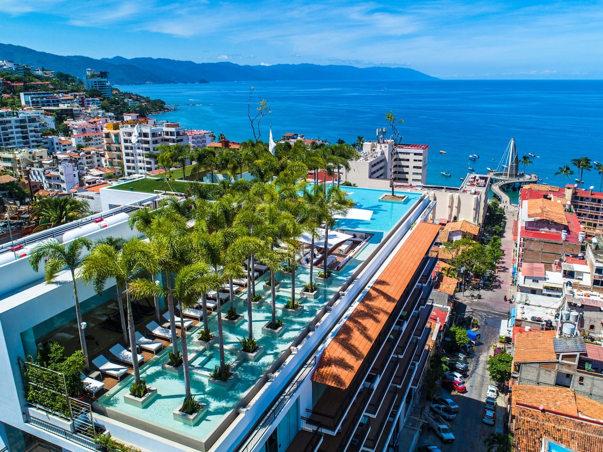 The rooftop terrace offers a panoramic view of Banderas Bay and the Sierra Madre Mountains. Lush palm trees are arranged around an infinity pool, complemented by lounge chairs. The vibrant city and coastline are visible in the distance, showcasing a harmonious blend of nature and urban life.