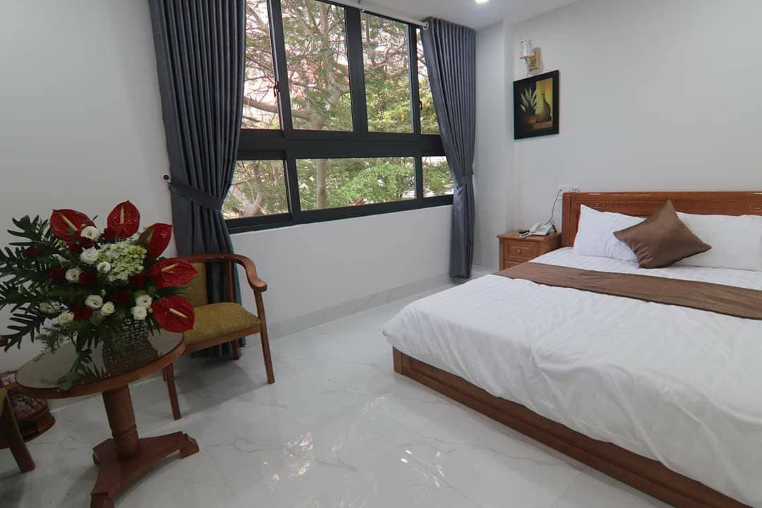 A spacious bedroom features a large bed with a neutral-colored blanket and decorative cushion, complemented by natural light from large windows. A small table with a chair and a floral arrangement add a touch of elegance to the inviting space.