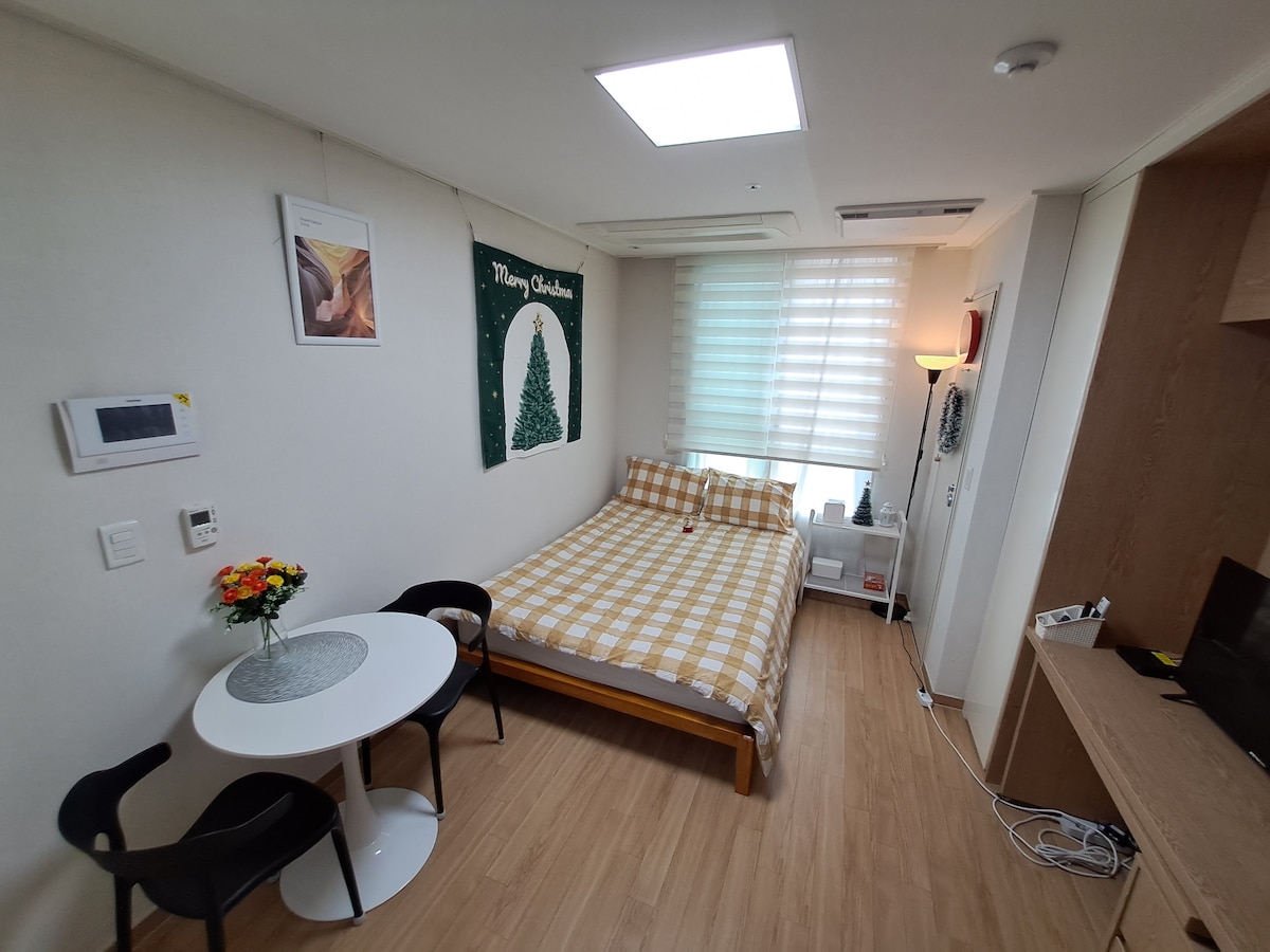 A bright room features a spacious bed adorned with a checkered blanket. A small round table with two chairs occupies one corner, accompanied by a vase of flowers. Natural light filters through sheer window coverings, enhancing the simple yet clean design.