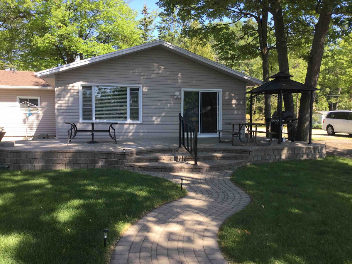 A well-maintained single-story house is set among greenery, featuring a paved path leading to a spacious stone patio. Outdoor seating is available under a gazebo, with additional tables and chairs positioned nearby. Large windows allow natural light to illuminate the interior.