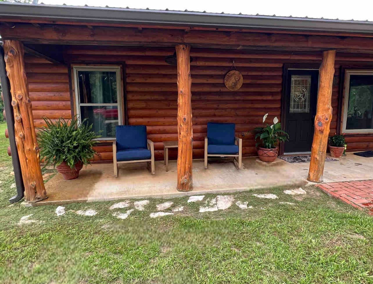 A rustic log cabin porch is showcased with two blue chairs positioned beside potted plants. The natural stone pathway leads to the entrance, while the sturdy wooden posts reflect the cabin's construction. Windows flanking the door allow for natural light.