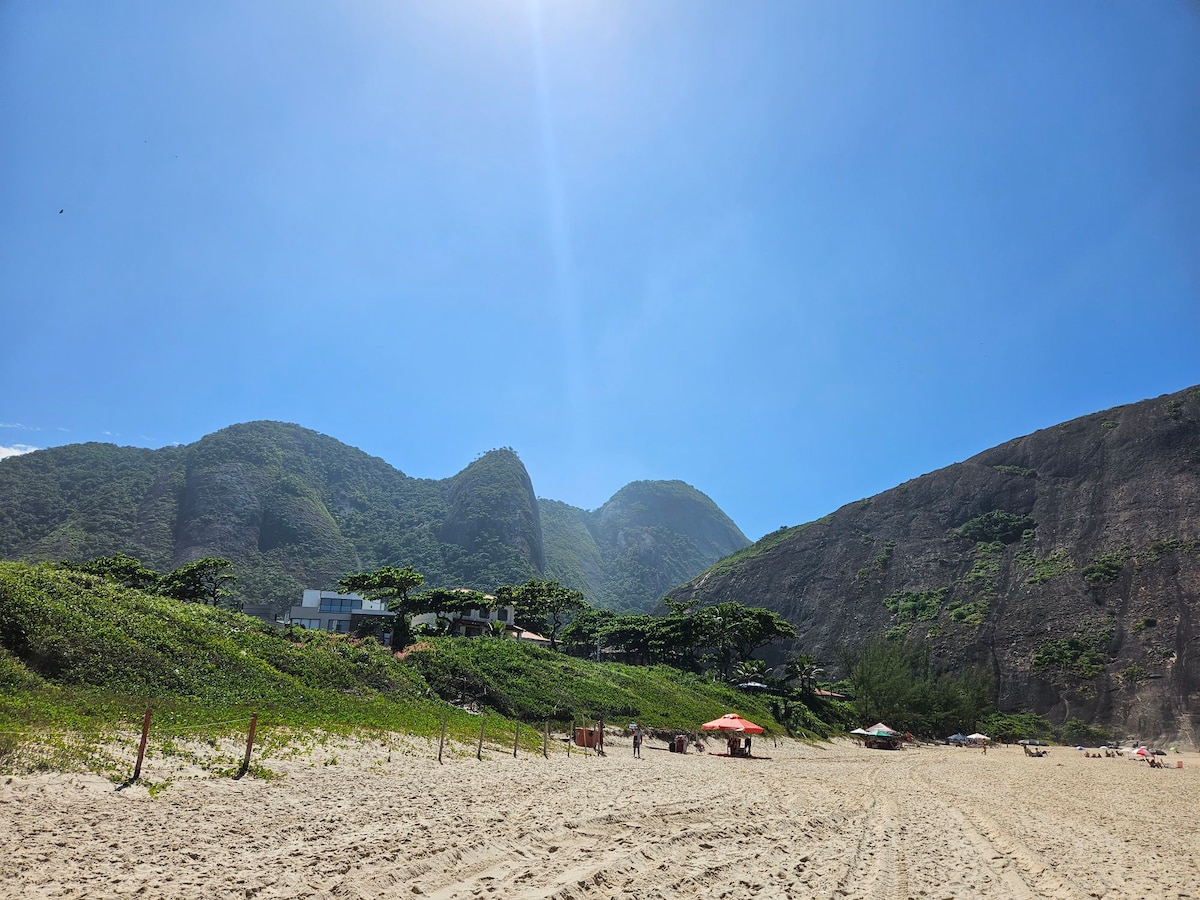 A sandy beach extends beneath a clear blue sky, bordered by green hills and mountains in the distance. Sun umbrellas are positioned along the shore, and a few visitors can be seen in the background, enjoying the peaceful natural surroundings.