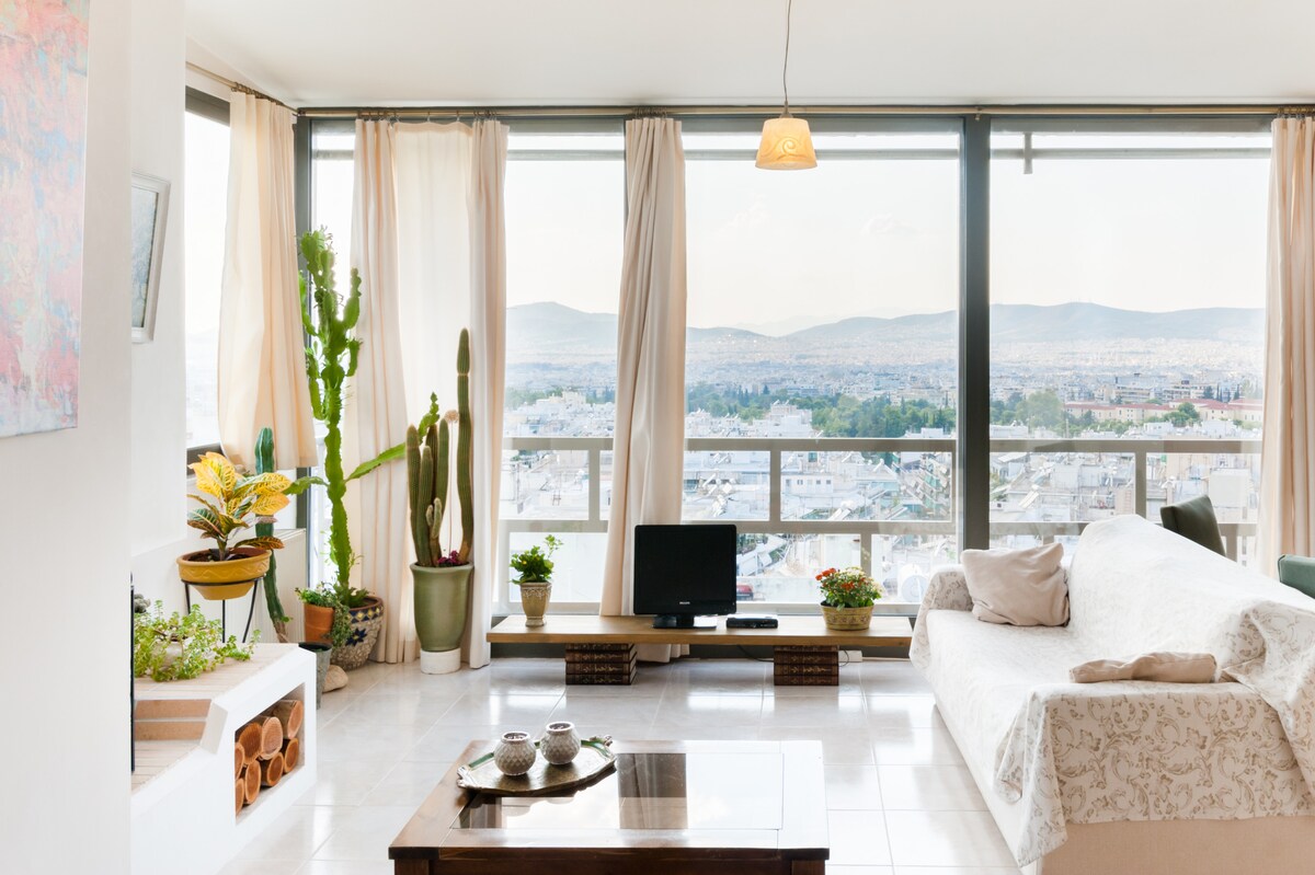A sunlit living area features large glass windows offering expansive views of the city and mountains. A white sofa is positioned beside a wooden coffee table. Various potted plants, including cacti, decorate the space, enhancing a vibrant ambiance. Natural light fills the room through sheer curtains.