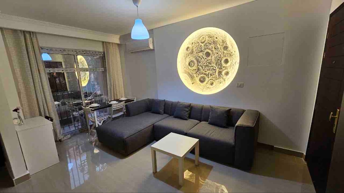 The living area features a spacious sectional sofa arranged around a minimalist white coffee table. A round, illuminated wall art piece draws the eye. Large windows allow for natural light and provide access to an outdoor seating area.