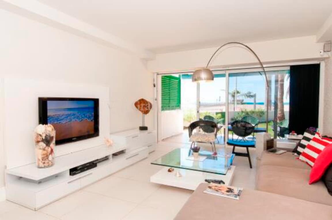 A modern living area features large glass doors that open to the outside, allowing natural light to fill the space. A flat-screen TV is mounted on a white entertainment unit, while a comfortable sofa adorned with red and white cushions invites relaxation. Stylish chairs surround a glass coffee table.